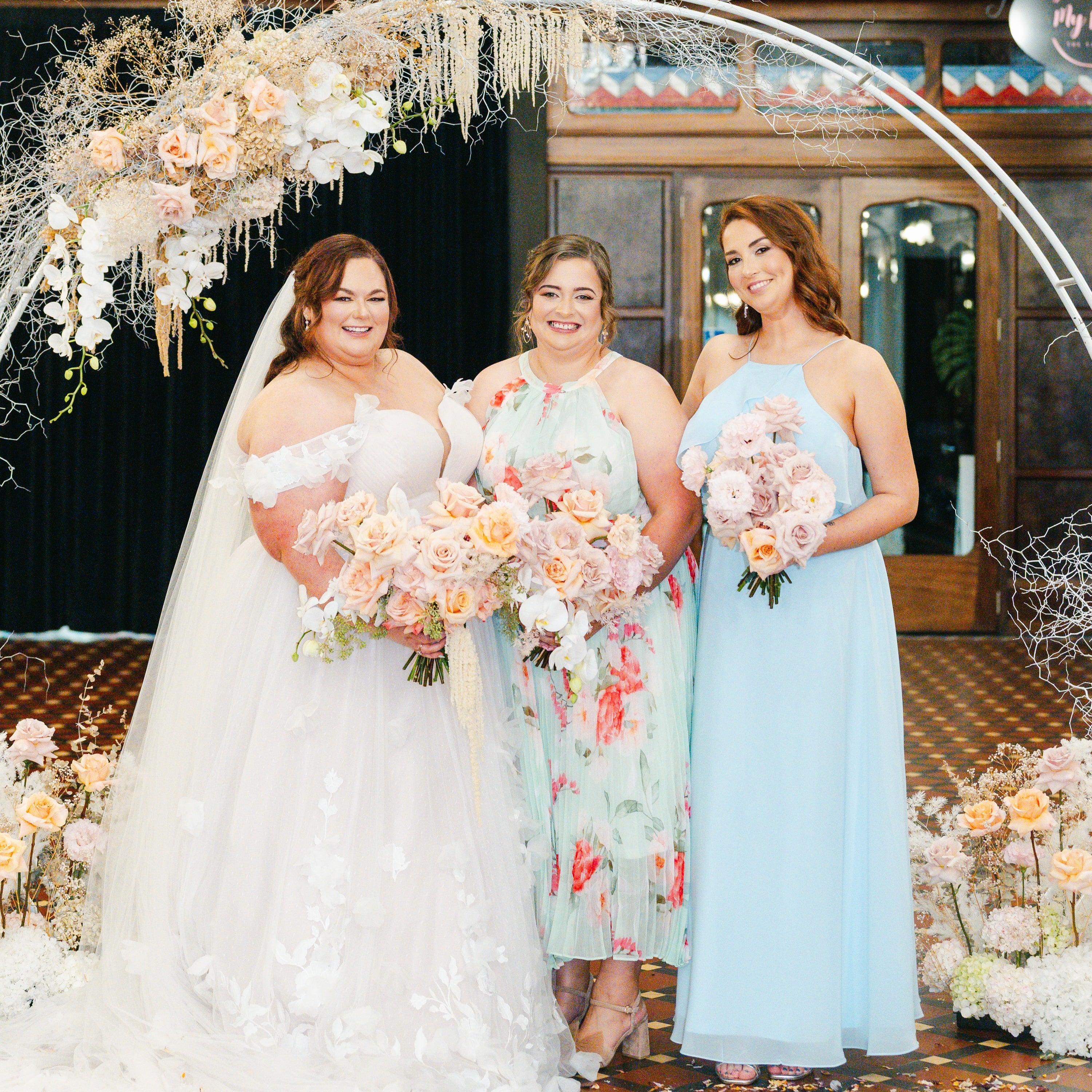 Three women standing under a floral archway with wedding bouquets, one in a white dress, another in a light blue dress, and the third in a floral dress.